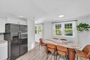 Eine moderne Küche mit einem Edelstahl-Kühlschrank, einem Marmortisch mit sechs Stühlen und einem Fenster mit Blick auf Grünflächen.