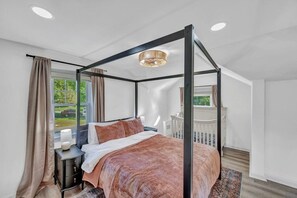 A four-poster bed with a canopy, a bedside table with a lamp, and a view of the outdoors through a window.