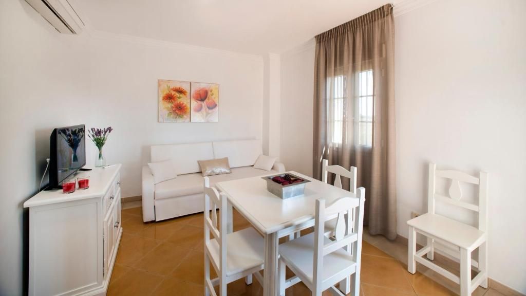 A white furnished room with a sofa, a dining table, chairs, a TV, and a window with curtains.