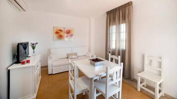 A white furnished room with a sofa, a dining table, chairs, a TV, and a window with curtains.