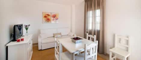 A white furnished room with a sofa, a dining table, chairs, a TV, and a window with curtains.