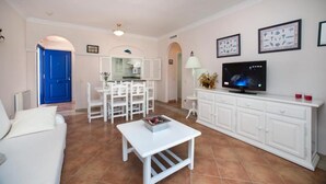 Una sala de estar con un sofá blanco, una mesa de centro blanca, un televisor y un mueble blanco.