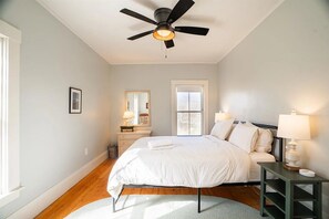 A bedroom with a bed, bedside tables, a ceiling fan, a window, and a door. - Riverfront 1800s Farmhouse, Sleeps 12+, Minutes from Killington + Woodstock (Bridgewater)
