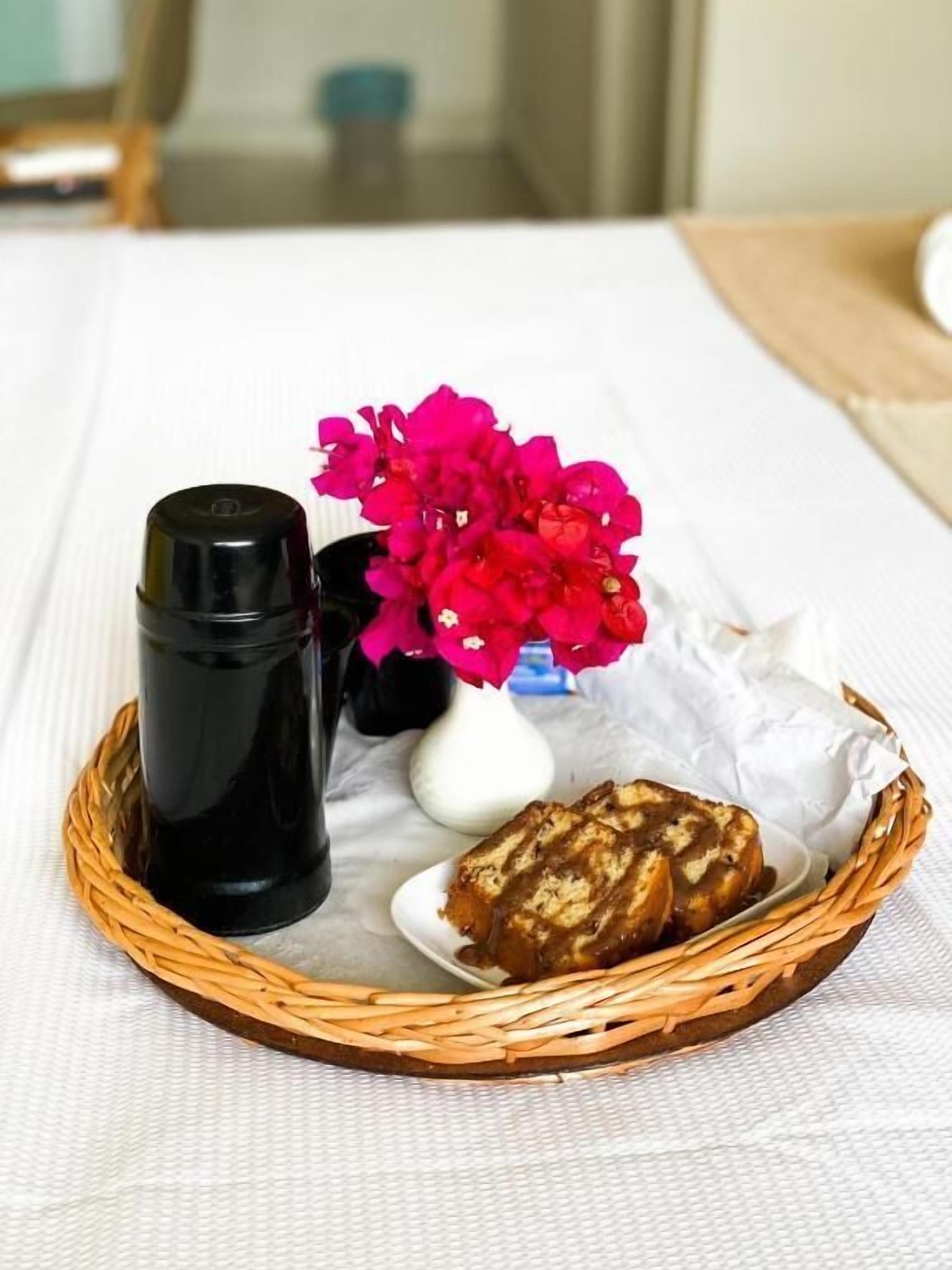 Uma mesa branca com uma cesta de vime contendo uma garrafa preta, um vaso branco com flores cor-de-rosa e um pedaço de pão.