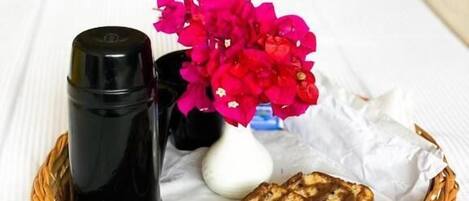 Uma mesa branca com uma cesta de vime contendo uma garrafa preta, um vaso branco com flores cor-de-rosa e um pedaço de pão.