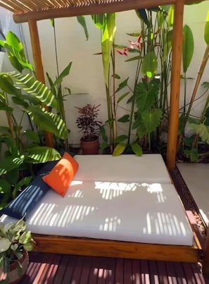 A wooden deck with a white mattress and orange pillow, surrounded by lush green plants. - Pousada Praiana (Ipojuca)