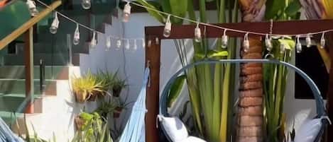 A hammock chair suspended from a wooden structure, surrounded by potted plants and a palm tree.