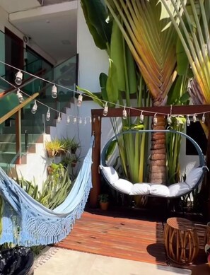 Una silla hamaca colgada de una estructura de madera, rodeada de macetas con plantas y una palmera.
