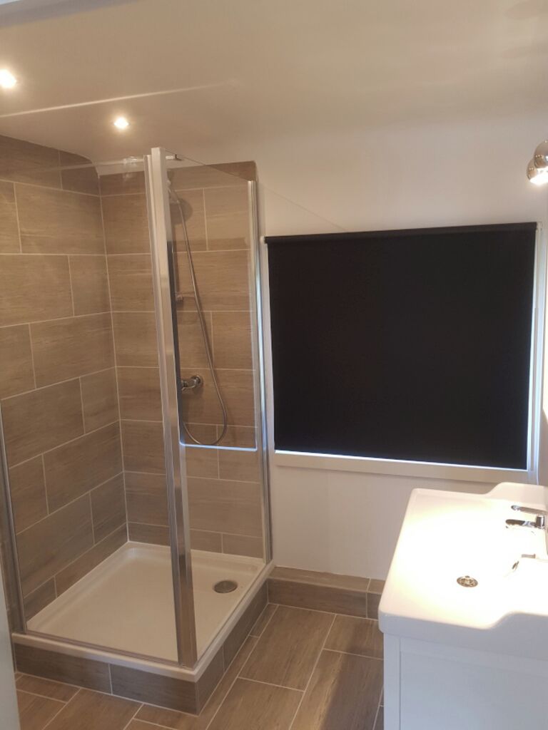 Une salle de bain avec une douche à parois de verre, un lavabo blanc et une fenêtre avec un store noir.