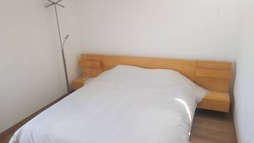 A single bed with a wooden headboard and a white mattress in a room with plain white walls and a wooden floor.