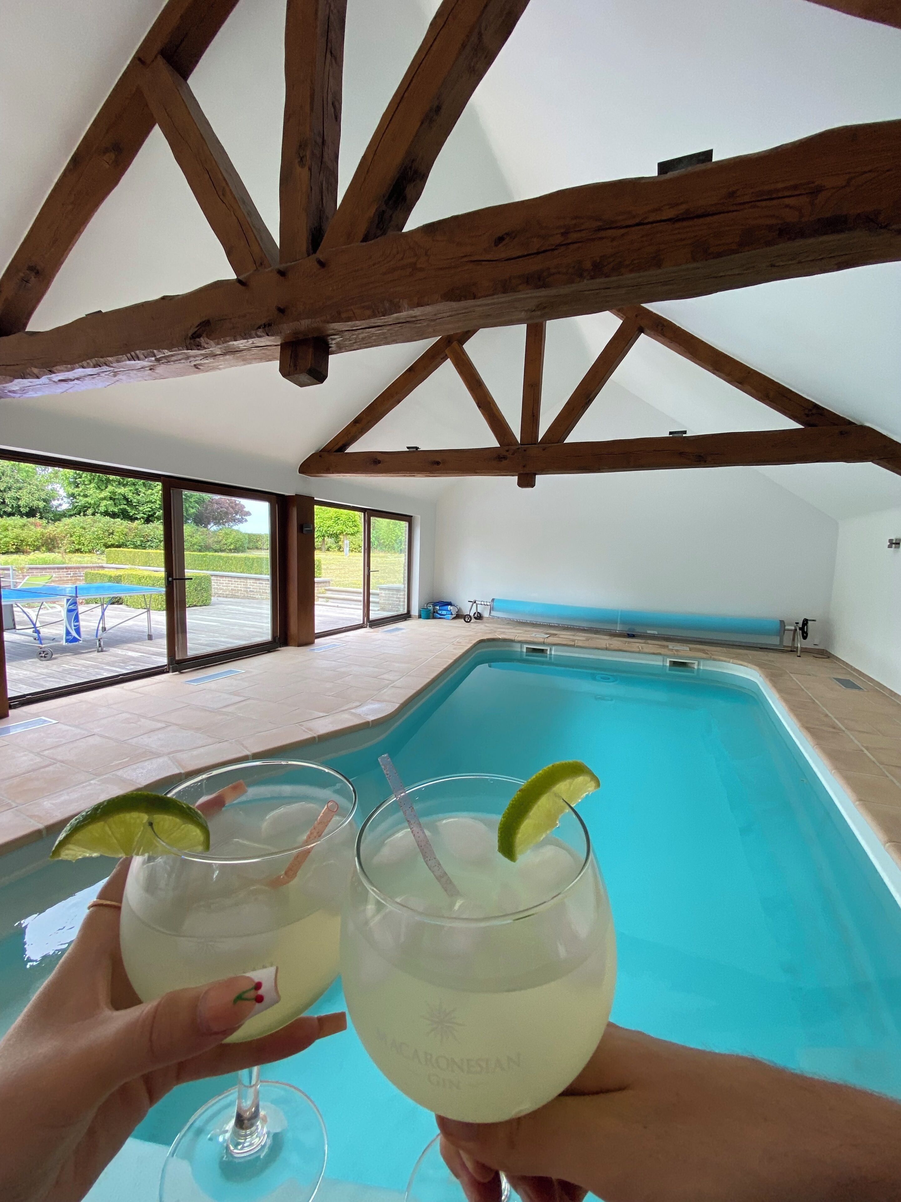 A pool area with a clear view of the outdoors, wooden beams on the ceiling, and two glasses of a clear liquid with lime slices.