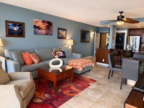Ein Wohnzimmer mit einer Couch, einem Couchtisch und einem Essbereich mit Stühlen. An den Wänden befinden sich Gemälde und es gibt einen Deckenventilator.