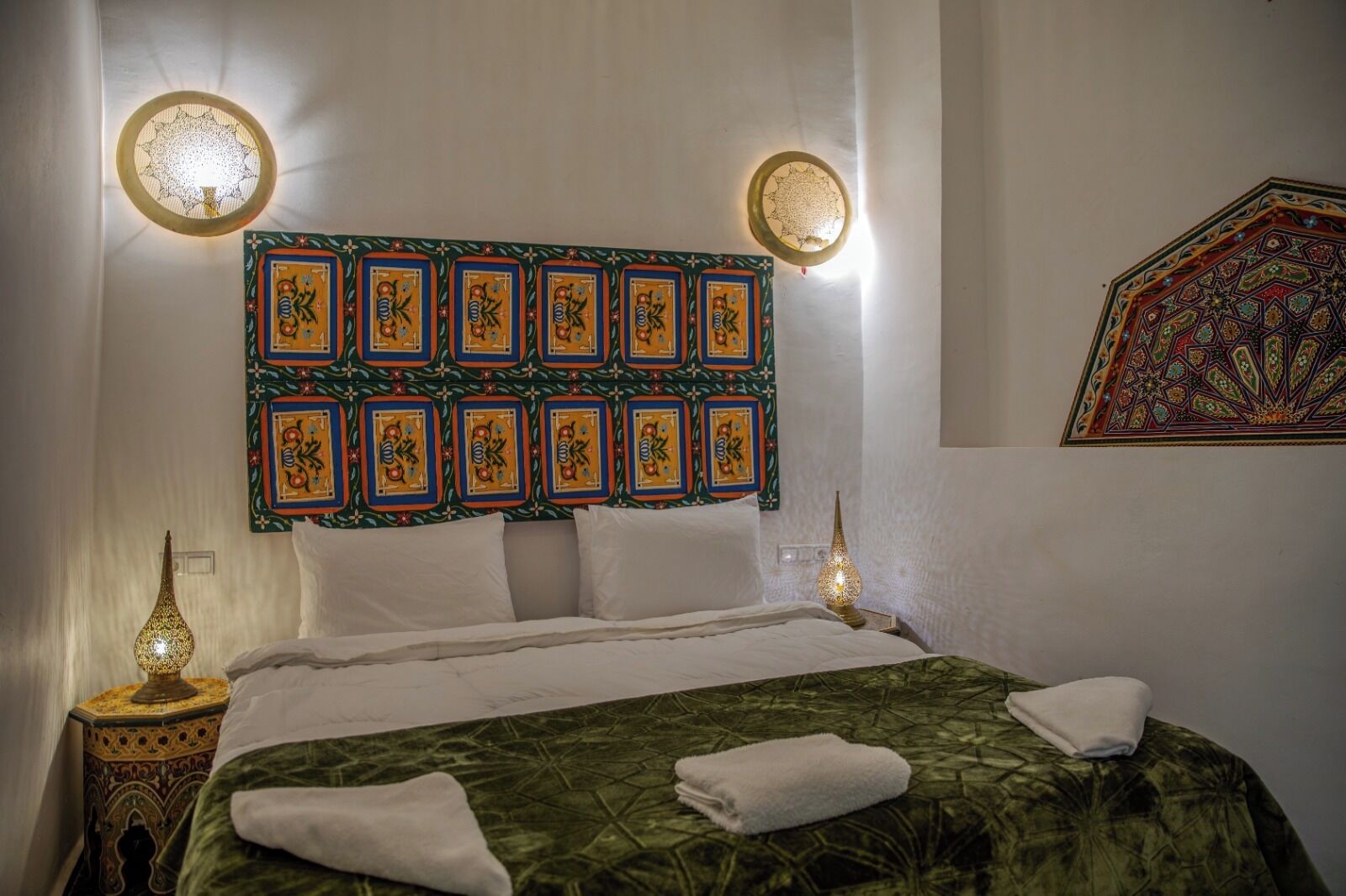 A bed with a patterned headboard, two wall-mounted lights, a decorative wall hanging, and a nightstand with a lamp.