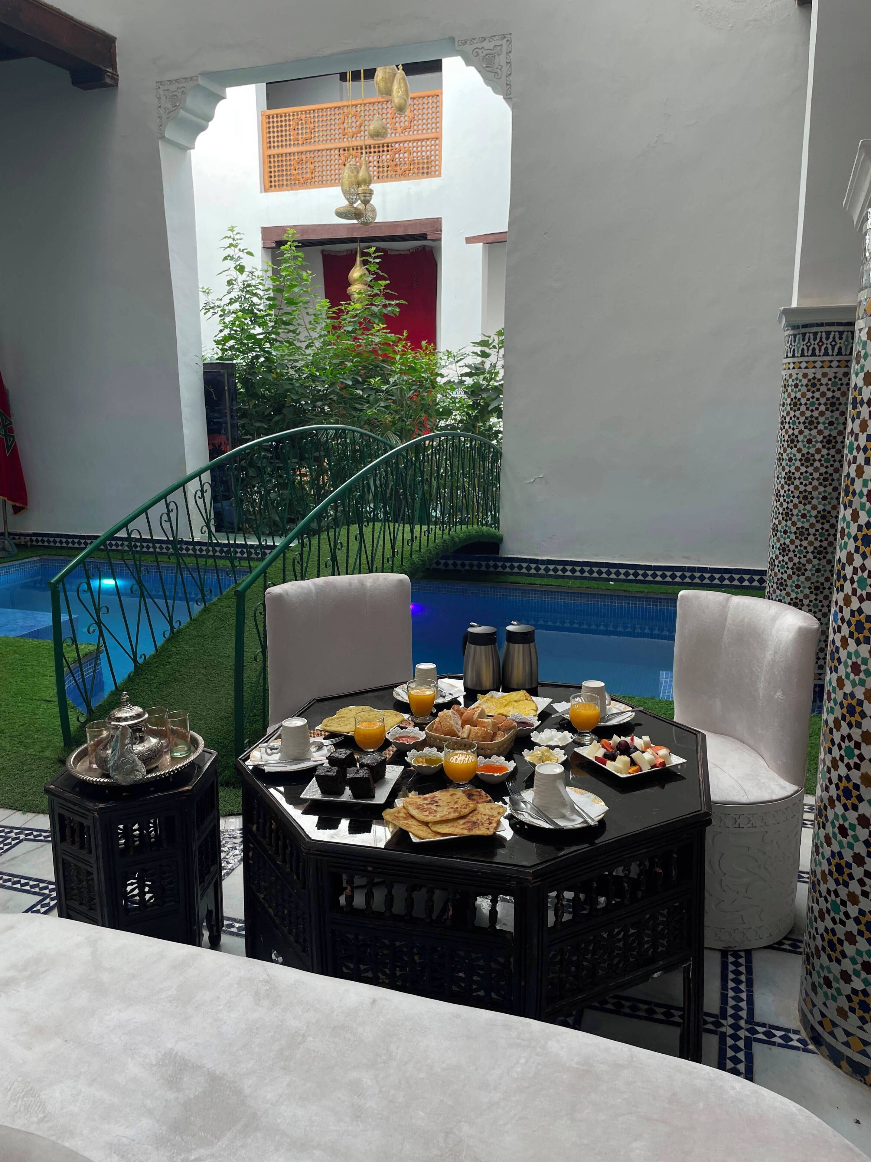 A dining area with a table set for breakfast, a pool in the background, and a staircase leading to a balcony.