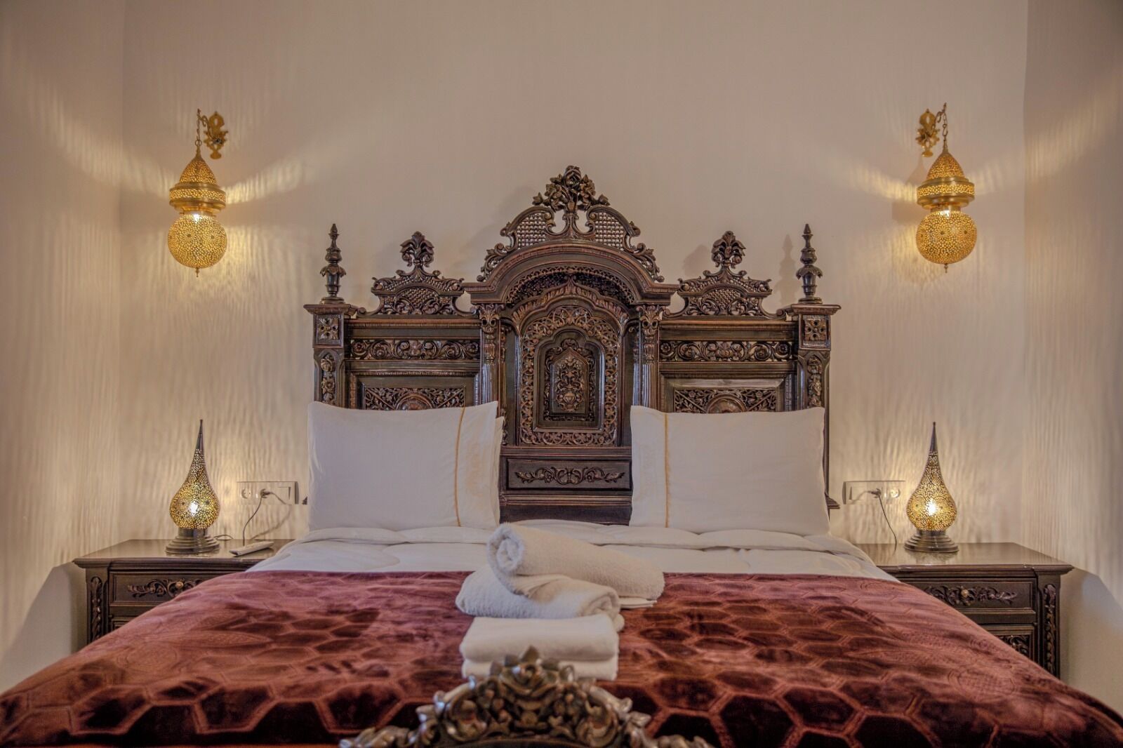 A bedroom with an ornate wooden headboard, two bedside lamps, and a red patterned bedspread.