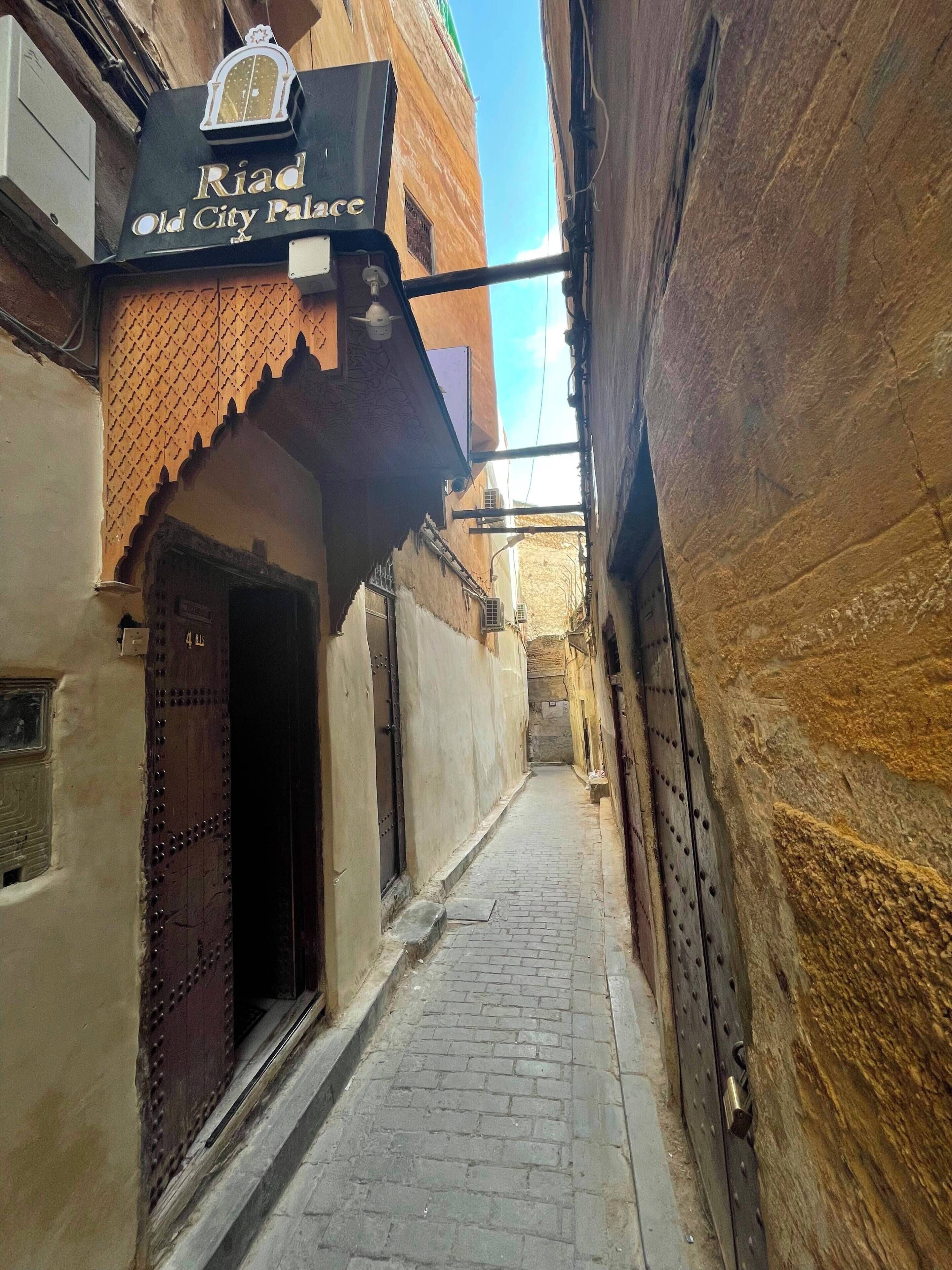 Een smalle steeg met een gebouw aan de linkerkant, voorzien van een bordje met de tekst 'Riad Old City Palace'.