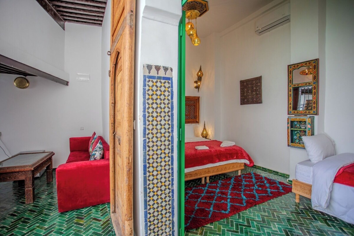 A room with a bed, a red sofa, a wooden table, and patterned floor tiles.