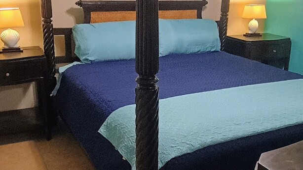 En säng med himmelstänger, blått sängöverkast och kuddar, med nattduksbord med lampor på var sin sida, i ett rum med en inramad tavla på väggen och en vägg i en turkos nyans som accentfärg.