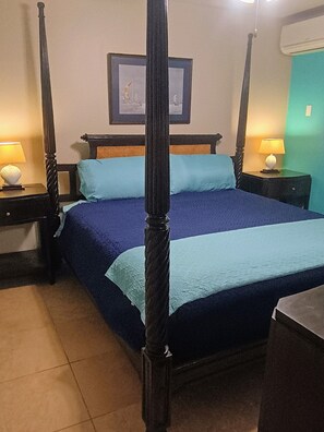 A four-poster bed with a blue bedspread and pillows, flanked by nightstands with lamps, in a room with a framed picture on the wall and a teal accent wall. - Vis Ton Reve- 2-bedroom condo in amazing Christiansted with AC and comfort (Christiansted)