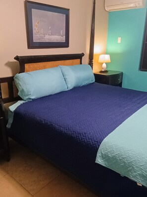 A bed with a blue quilt and pillows, a wooden headboard, a nightstand with a lamp, and a framed picture on the wall. - Vis Ton Reve- 2-bedroom condo in amazing Christiansted with AC and comfort (Christiansted)