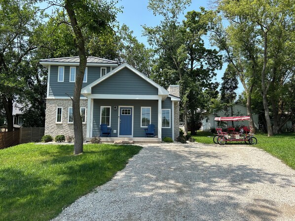 A two-story house with a front porch, a gravel driveway, and a red wagon in the background. - The Lake Escape! (Lake Geneva)