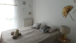 A bedroom with a bed, pillows, a rolled towel, a radiator, wall art, and a lamp.