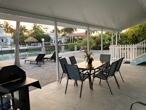 An outdoor seating area with a table and chairs, a grill, and a view of a water body and buildings in the background. - Waterfront Canal Priv Home. 3BD, 2BA, Pool, Kayak+Cabana Club! 70' Seawall-KCB (Marathon)