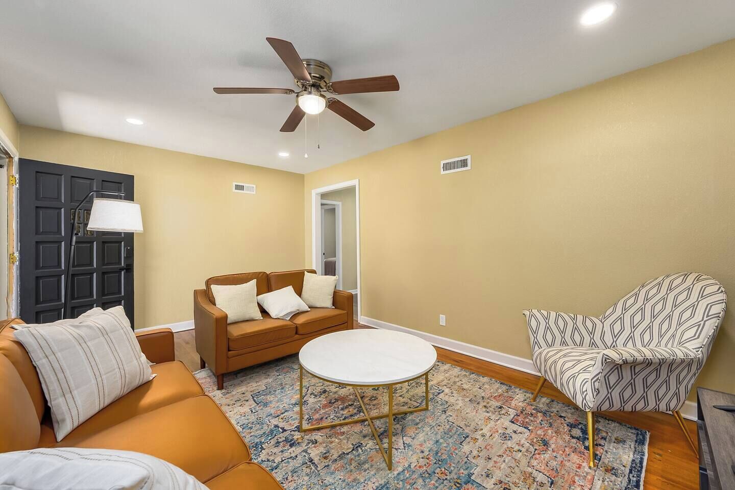 A living room with a brown sofa, a patterned armchair, a round coffee table, a ceiling fan, and a door with a black frame.