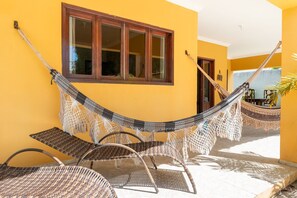 A hammock and wicker chairs in a sunny outdoor area with a yellow wall and a window. - Mansão-7quartos em Porto de Galinhas-165 (Camela)