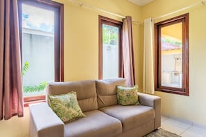 A beige sofa with two pillows, positioned in front of a window with brown curtains. - Mansão-7quartos em Porto de Galinhas-165 (Camela)