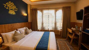 A hotel room with a bed, bedside table, lamp, TV, desk, chair, and curtains.