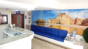 Uma área de recepção com um sofá azul, um balcão com tampo de vidro, um mural com paisagem de praia e placas indicando 'SQUASH', 'BALNEÁRIOS', 'TOILETS' e 'LAVANDERIA'.