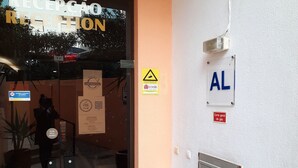 Uma área de recepção com porta de vidro, planta em vaso, lixeira e aviso de gás fixado na parede.