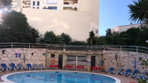Piscina com espreguiçadeiras, um muro de pedra e um edifício ao fundo.