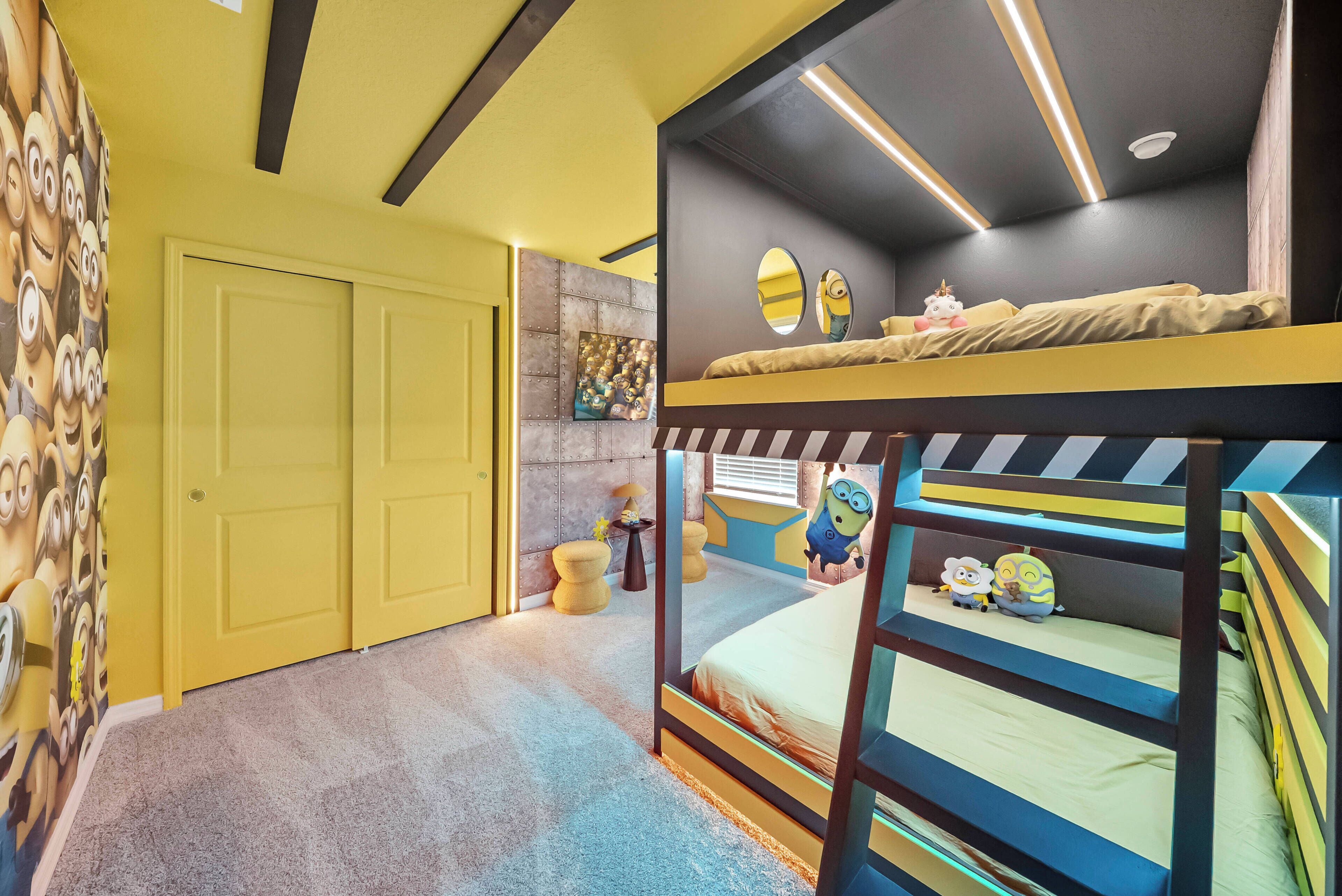 A room with bunk beds, a door, a wall with a Minion-themed mural, and a small table with a lamp.