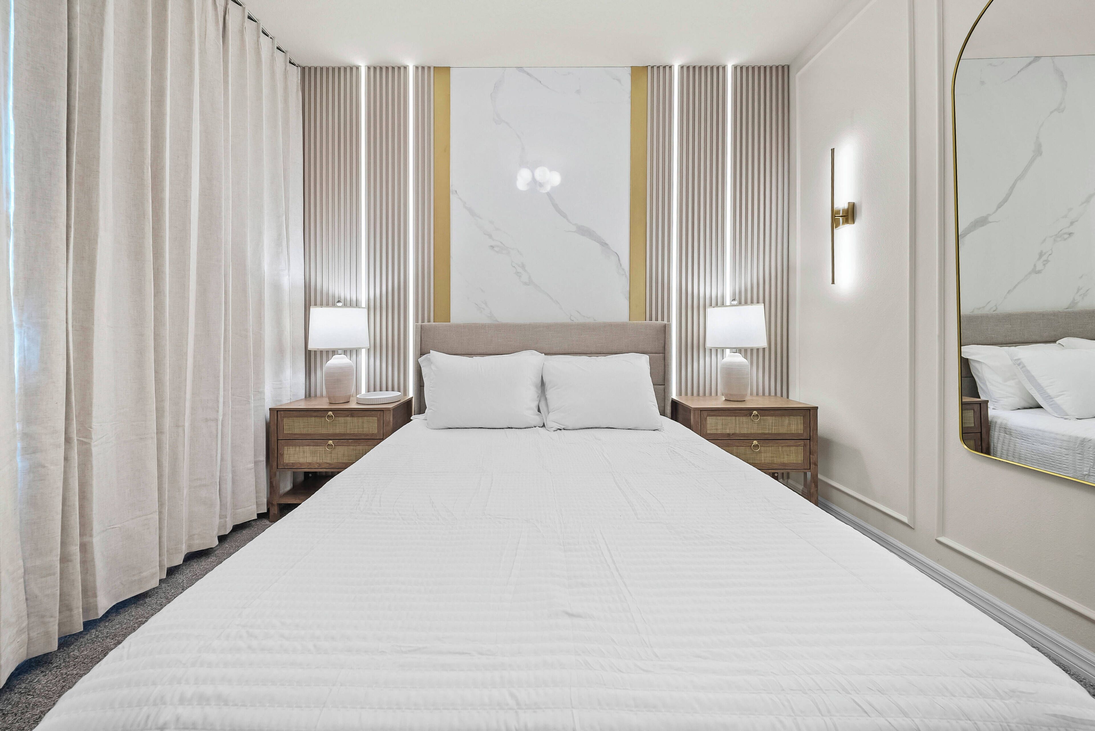 Quarto de hotel com uma cama grande, duas mesas de cabeceira com luminárias, um espelho e uma cortina listrada.