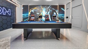 Uma mesa de bilhar com três máquinas de jogos eletrônicos, uma placa de neon e um luminário hexagonal no teto.