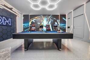 Ein Billardtisch, drei Spielautomaten, eine Neonreklame und eine sechseckige Leuchte an der Decke.