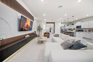 A modern living room with a flat-screen TV, a sofa, a coffee table, and a kitchen area with white cabinets and a microwave. - 8BR house with private pool and game room (Clermont)