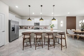 Une cuisine moderne dotée d’un îlot central, de tabourets de bar, d’appareils électroménagers en acier inoxydable, de luminaires suspendus et d’un coin repas avec une table et des chaises.