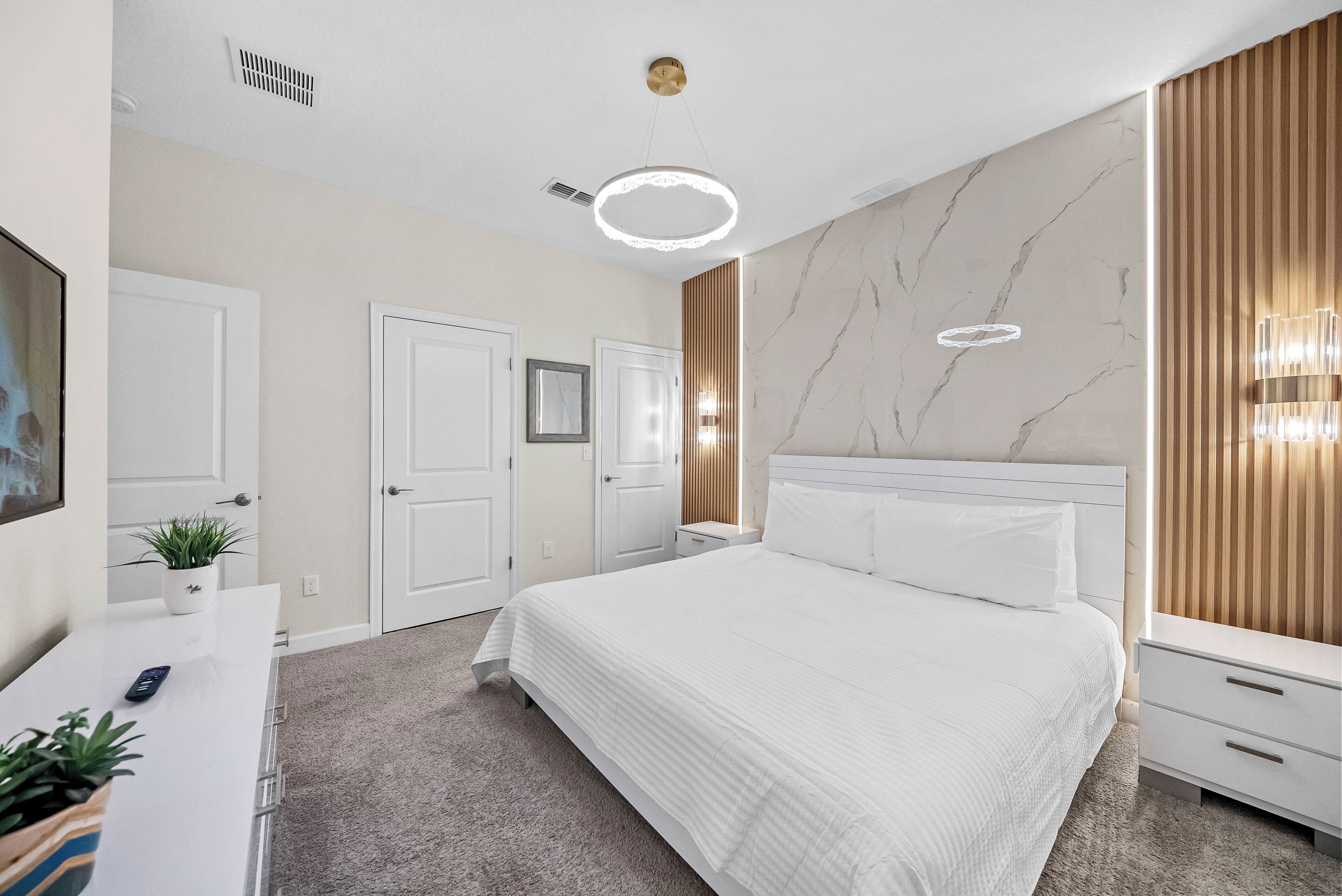 A modern bedroom with a large bed, a bedside table, a nightstand, a wall-mounted light fixture, and a ceiling vent.
