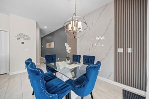Een moderne eethoek met een glazen tafel, blauwe fauteuils, een kroonluchter en een marmeren wand.