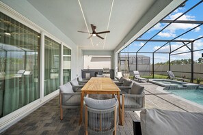 A modern outdoor patio with a wooden table and chairs, a ceiling fan, and a pool area with lounge chairs. - 10BR house at Windsor Cay Resort in florida (Clermont)