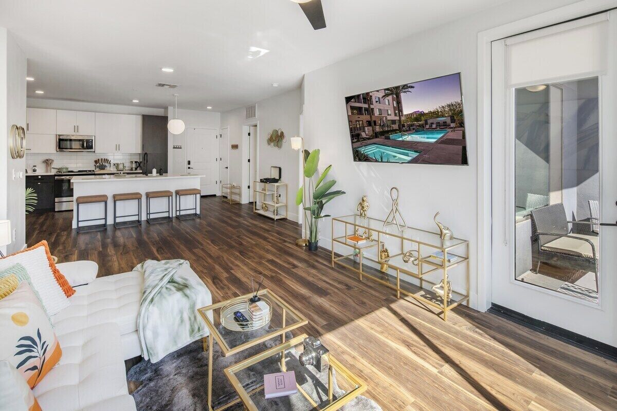 A modern living room with a sofa, coffee table, and a dining area with a bar. There is a large window with a view of a swimming pool.