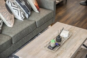 Ein modernes Wohnzimmer mit einem grauen Sofa, verziert mit gemusterten Kissen, einem Couchtisch aus Holz mit einer Ablage für eine kleine Pflanze, eine Kerze und einen Diffusor.