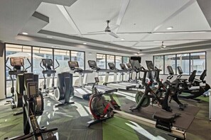 Ein Fitnessstudio mit verschiedenen Trainingsgeräten, großen Fenstern und einem Deckenventilator.