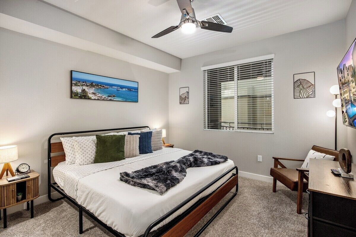 A bedroom with a bed, a ceiling fan, a window with blinds, a bedside table with a lamp, and a wall-mounted TV.