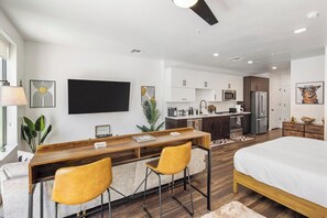 A modern bedroom with a large bed, a wooden desk with two chairs, a kitchenette, and a TV mounted on the wall. - Escape To Poolside Bohemian Desert King Bed Studio (Tempe)