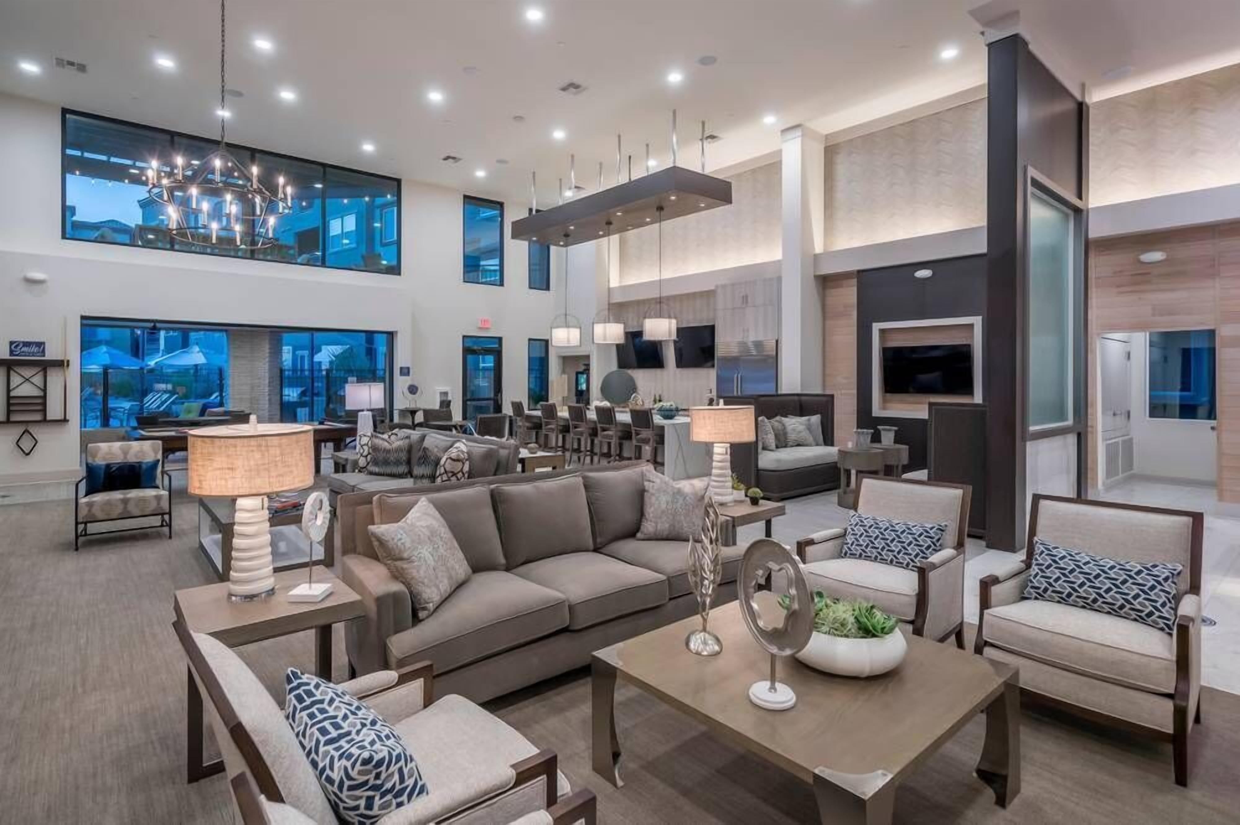 A spacious living area with a large sofa, armchairs, a coffee table, and a dining table. There are multiple ceiling lights and a large window providing a view of the outdoors.