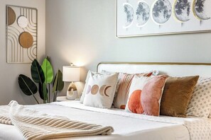 A neatly made bed with a white comforter and a beige throw blanket, a bedside table with a lamp, and a wall with abstract art and a framed picture of moons. - Escape To Hollywood Hideaway 2BR King Bed Suite (Phoenix)